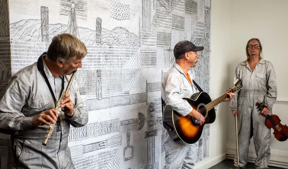 An image of three people playing musical instruments in front of a large scale drawing