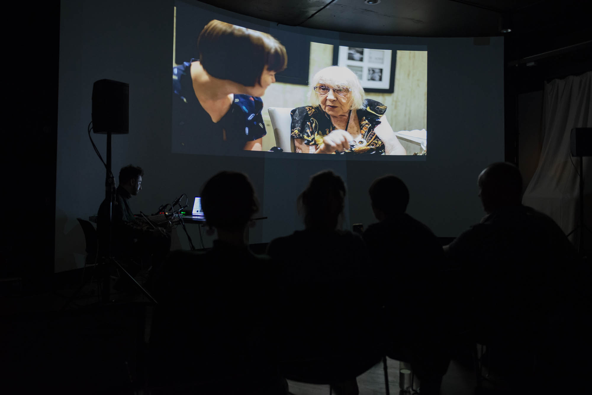 A photograph of a darkened space with the silhouettes of audience visible and the artist on screen speaking with an elderly woman.