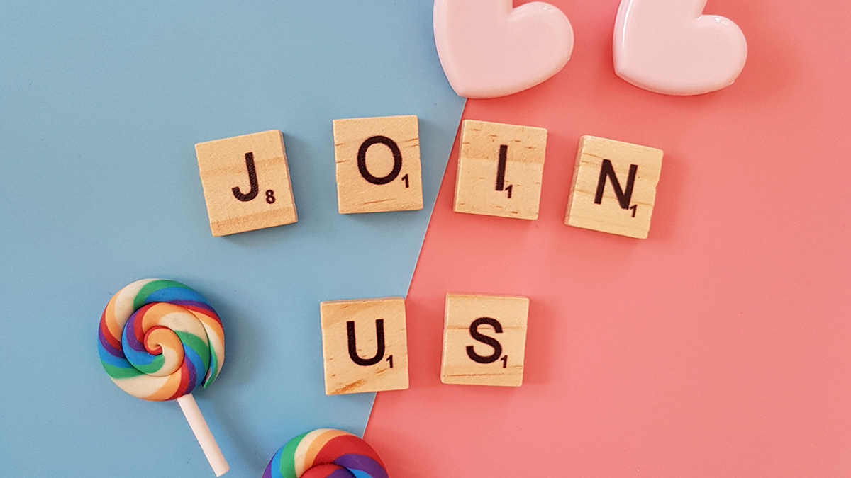 The words 'Join Us' written in Scrabble tiles on a pastel pink and blue background, with sweets