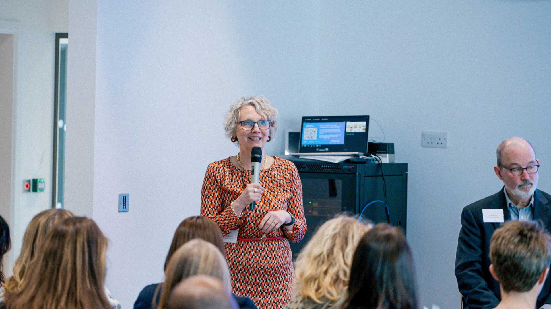Dr Nic Stenberg standing in front of a group of people, holding a microphone and smiling.
