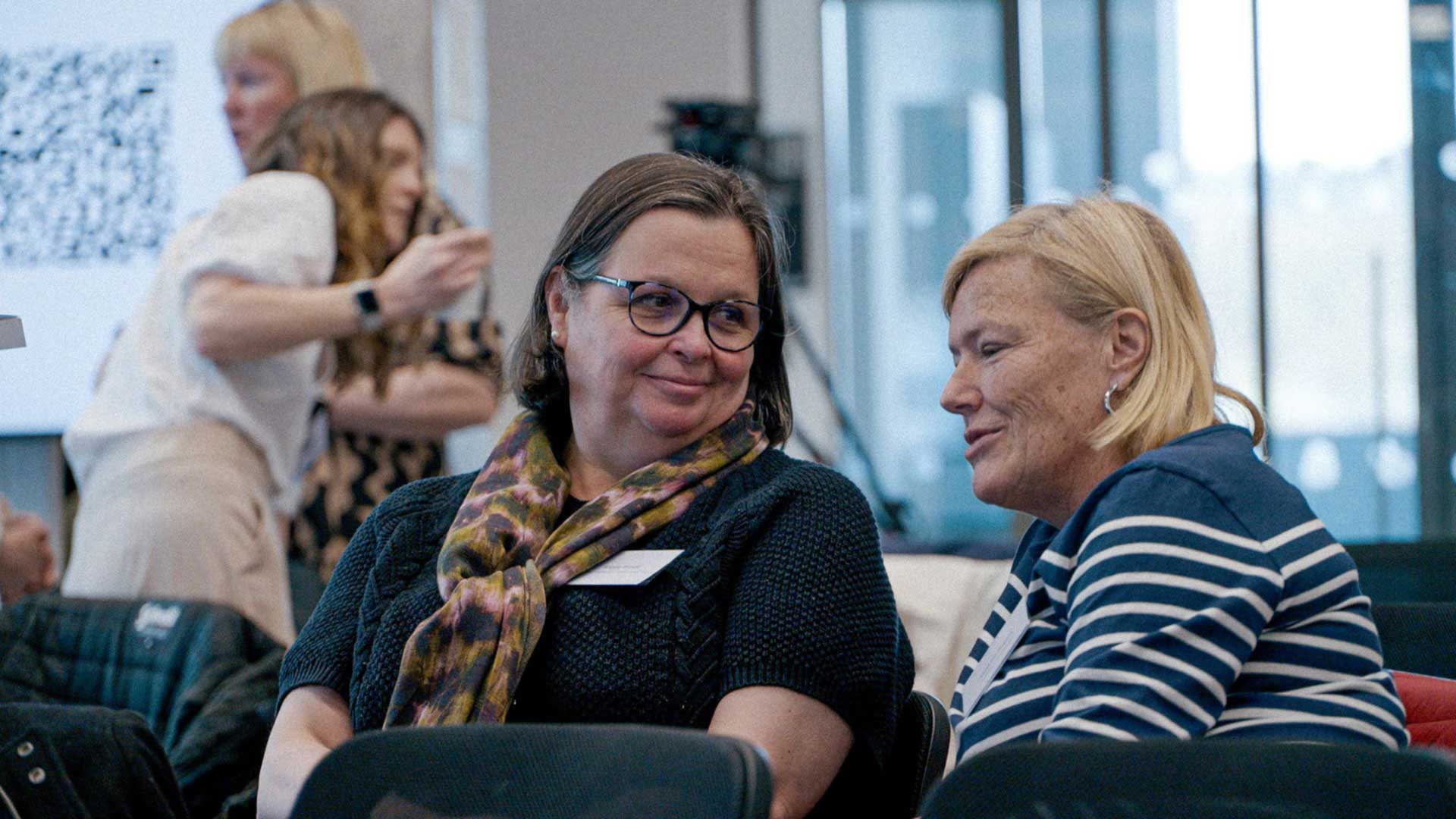 Two people sat on a couch, looking at eachother and smiling.